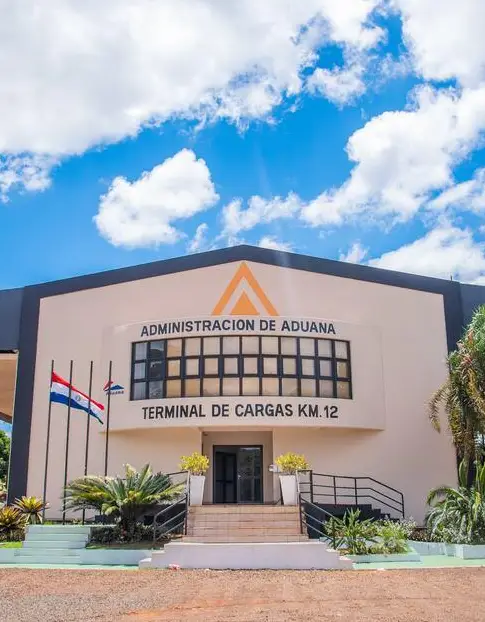 Het gebouw van de Administracion de Aduana Terminal de Cargas Km. 12 onder een blauwe hemel. Links van de ingang, die via een trap bereikbaar is, wappert de Paraguayaanse vlag.