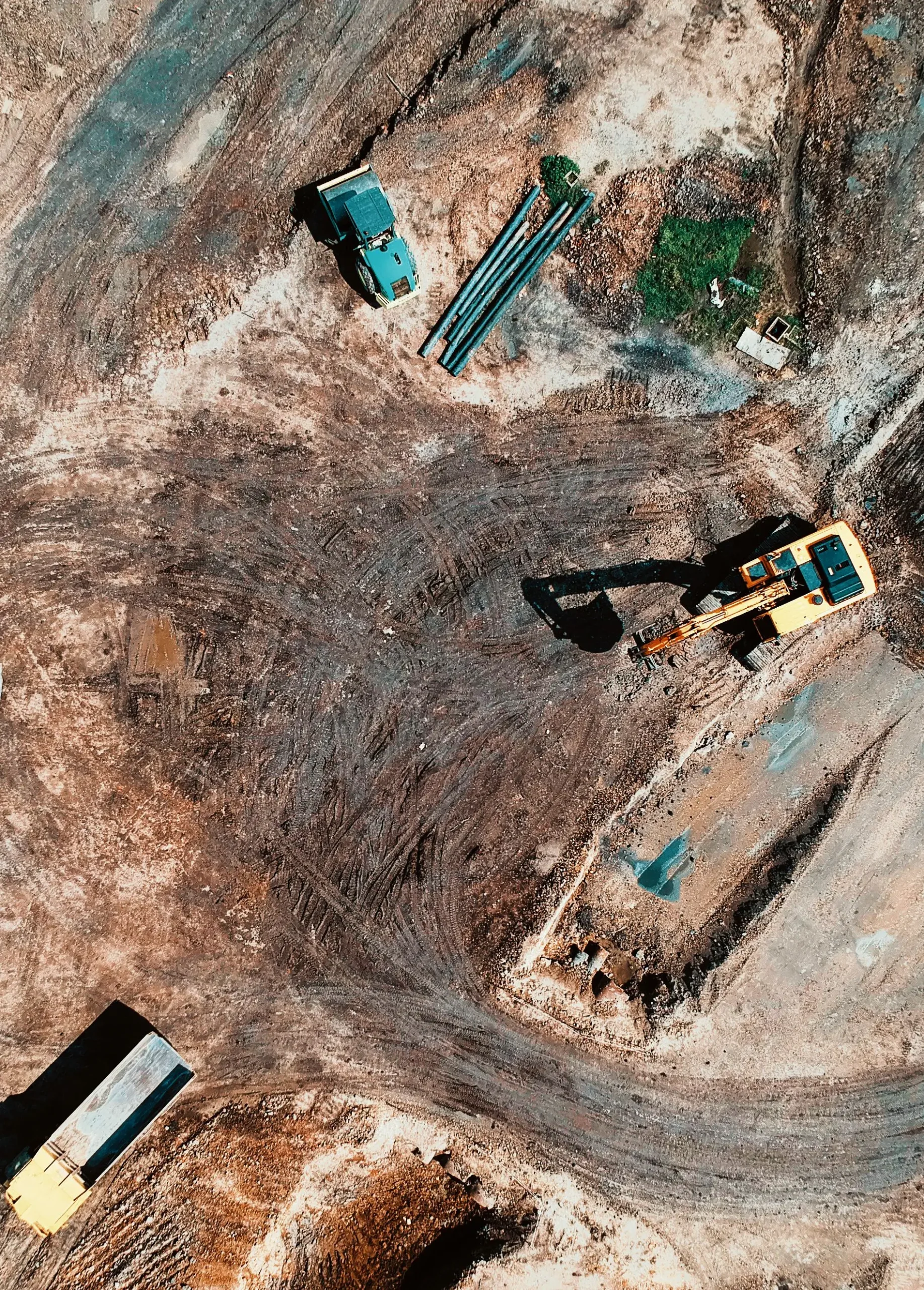 Luchtfoto van een bouwplaats met een gele graafmachine, een blauwe wals en een kiepwagen op een onverharde ondergrond. Tussen de voertuigen liggen drie metalen buizen.