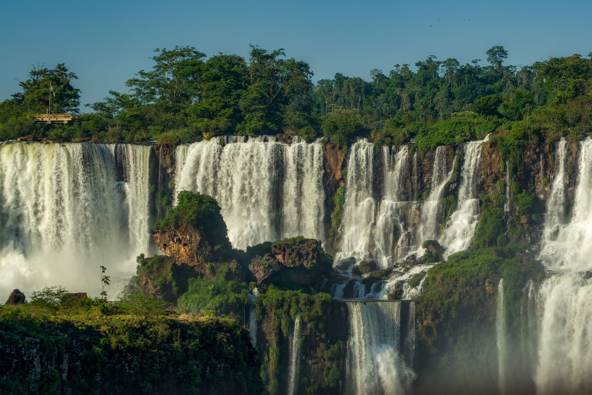 Verschillende watervallen stromen over een rotsachtige jungle langs de klif, te zien vanaf een bergkam die wellicht in de smaak valt bij mensen die Visaloket Paraguay willen bezoeken. Links op de met bomen omzoomde bergkam bevindt zich een klein uitkijkplatform.