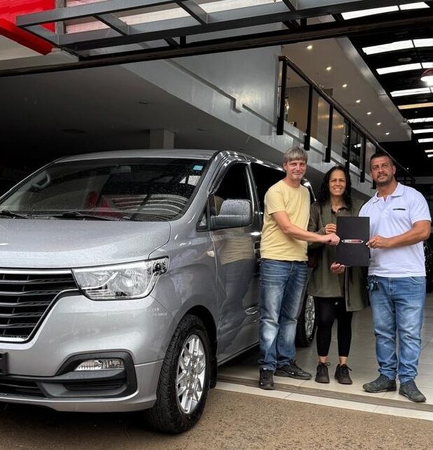 Twee mannen en een vrouw staan naast een nieuw zilverkleurig busje bij een autodealer. Ze houden samen een map vast en vieren de aankoop van het voertuig.