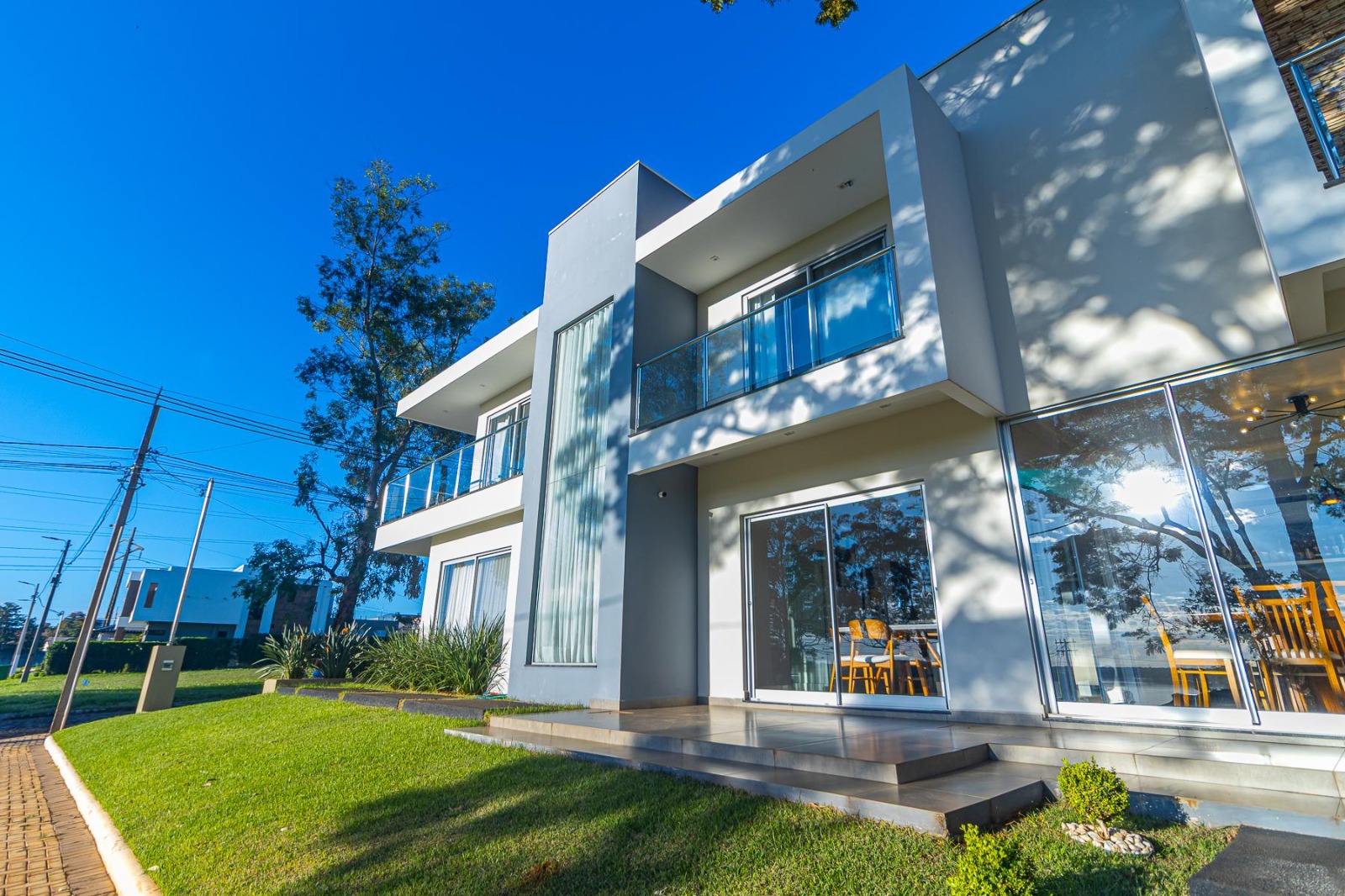 Moderne woning met twee verdiepingen, witte muren en glazen balkons. De kamerhoge ramen kijken uit op een eethoek, en aan de voorzijde strekt zich een groen gazon uit – ideaal voor wie ervoor kiest om succesvol te emigreren via Visaloket Paraguay.