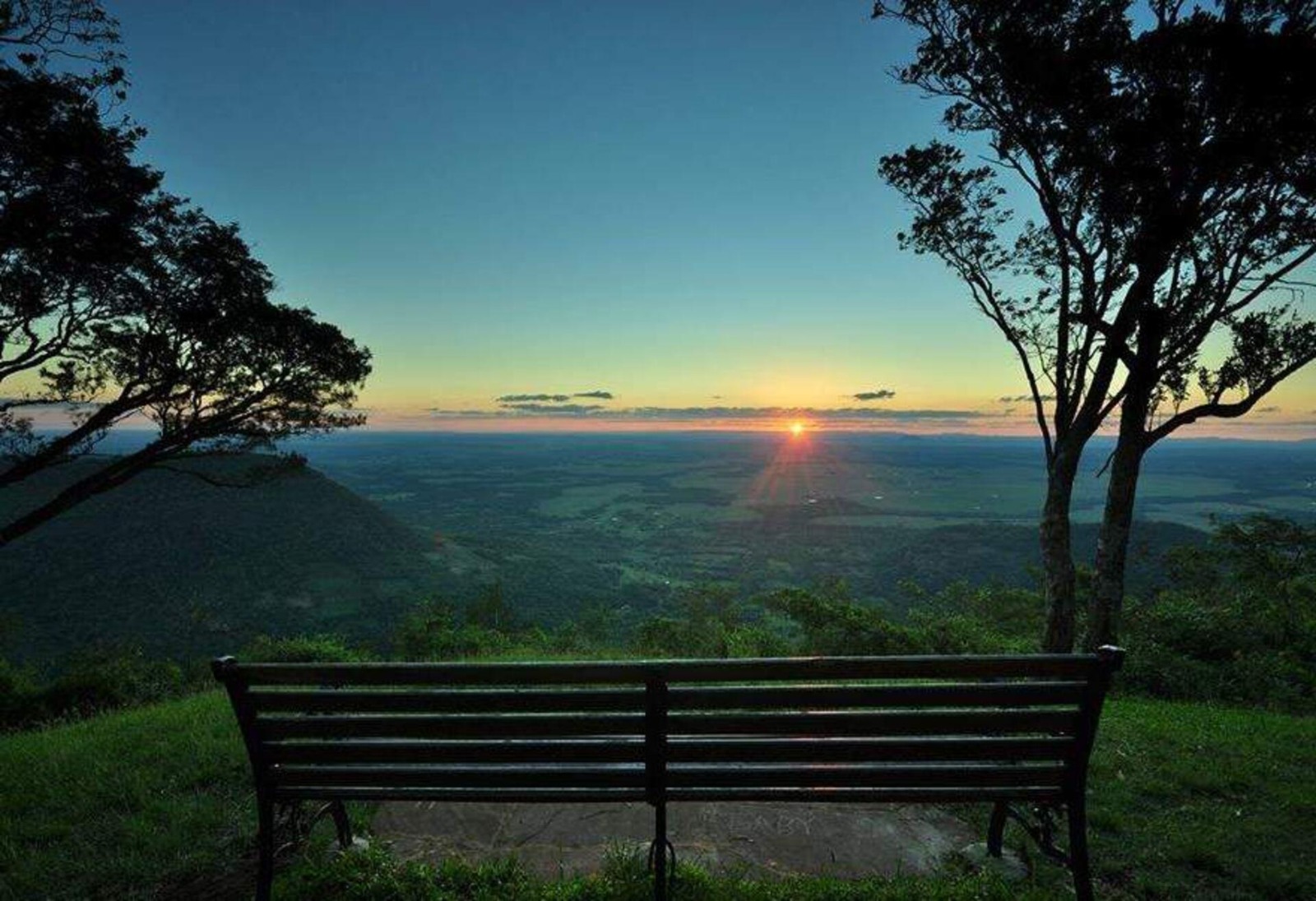 Op een met gras begroeid uitkijkpunt staat een metalen bankje, vanwaar je een uitzicht hebt op een zonsondergang die het gevoel van een succesvolle emigratie met Visaloket Paraguay perfect weergeeft. Bomen omlijsten het uitgestrekte landschap aan weerszijden van de horizon.